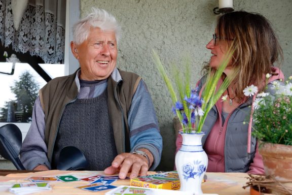 Alltagsbegleitung mit Herz Älterer Mann und Frau im Gespräch, umgeben von Pflanzen und Karten auf dem Tisch.