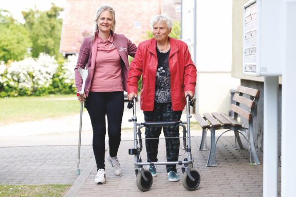 Alltagsbegleitung mit Herz Zwei Frauen gehen gemeinsam, eine mit einem Rollator, in einer hellen Umgebung.