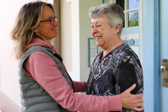 Alltagsbegleitung mit Herz Zwei Frauen umarmen sich lächelnd vor einem Gebäude.