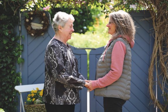 Alltagsbegleitung mit Herz Zwei Frauen stehen sich gegenüber und halten sich an den Händen vor einem blauen Tor.
