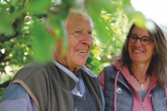 Alltagsbegleitung mit Herz Älterer Mann und Frau stehen unter Bäumen, lächeln und genießen die Zeit zusammen.