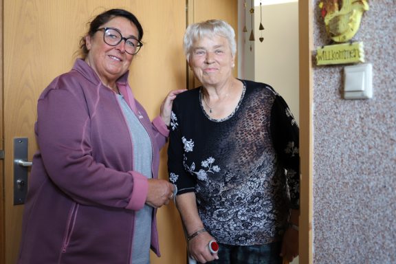 Alltagsbegleitung mit Herz Zwei Damen stehen lächelnd vor einer Tür, eine hält einen Gehstock.