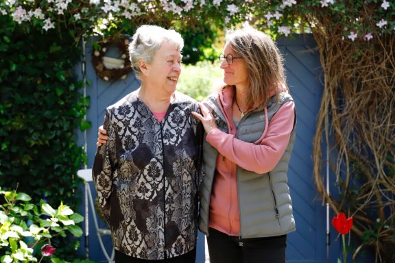 Alltagsbegleitung mit Herz Zwei Frauen lächelnd im Garten, eine ältere Frau und eine jüngere, in enger Umarmung.